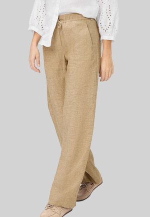 Vrouw die beige linnen wijde pijpenbroek draagt, wit eyeletblouse en beige veterschoenen, staande tegen een effen grijze achtergrond.