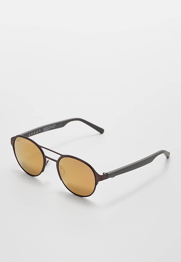Gafas de sol redondas con lentes marrones en degradado, montura de metal oscuro y brazos texturizados en negro. Presenta un logotipo mínimo en la montura.