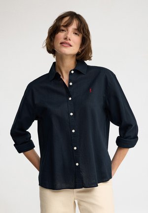 Kvinde med kort brunt hår iført en mørkeblå skjorte med knapper og opkrogede ærmer og lyse bukser, står med hænderne bag ryggen.