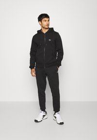 Černá mikina na zip s kapucí a pružnými šňůrkami, doplněná o odpovídající černé tepláky. Bílé sneakers s černými detaily.