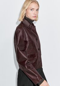 Vinrød læderjakke med krave, to knapper foran og en sidelomme. Glat tekstur og skræddersyet pasform.