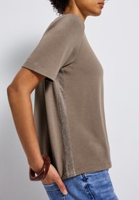 Hnědý tričko s krátkým rukávem a texturovaným kovovým bočním pruhem. Měkká látka, volný střih a jednoduchý design. Nosené s džíny.