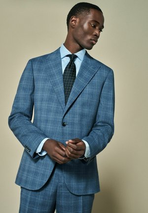 Abito a quadri blu con rever a scasso, chiusura a un bottone e cravatta nera su una camicia azzurra chiara. Tessuto strutturato con vestibilità sartoriale.