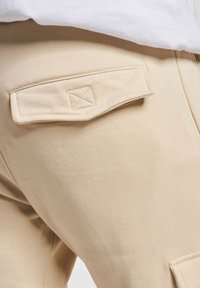 Beżowe spodnie cargo z naszywaną kieszenią z detalem w formie krzyżowego ściegu, wykonane z miękkiego, teksturowanego materiału, odzwierciedlające swobodny styl.