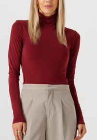 Bordeaux lange mouwen turtleneck top van rekbare stof, gecombineerd met lichtbeige high-waisted broek met voorplooien.