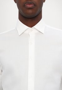 Camisa branca de dress shirt com colarinho padrão, apresentando uma frente com botões e uma textura subtil. Logo bordado no lado esquerdo do peito.