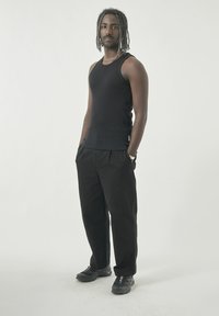 Schwarzes geripptes Tanktop, schwarze locker sitzende Hose und schwarze Sportschuhe. Akzent mit Silberkette. Die Person steht mit den Händen in den Taschen.