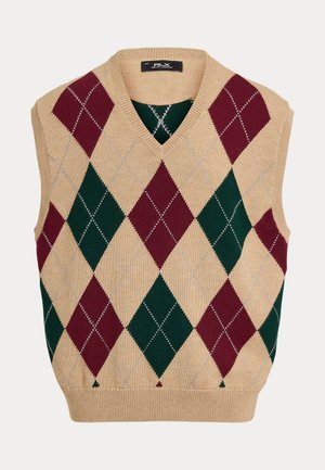 Homme portant un gilet en laine à losanges vert et bordeaux sur une chemise blanche, pantalon gris, chaussures blanches, casquette verte et lunettes de soleil, debout devant un fond beige.
