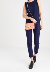 Granatowy top bez rękawów, granatowe spodnie o dopasowanym kroju i czarne wsuwane buty; trzyma małą koralową torebkę na ramię z metaliczną klamrą.