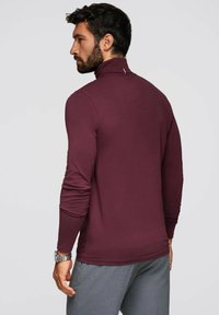 Kastanjefärgad långärmad skjorta med hög krage, tillverkad av mjukt tyg, med en diskret logotypdetalj på baksidan. Stylistiskt ihop med gråbyxor.