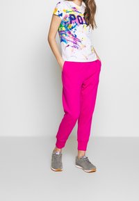 Bílá tričko s barevnými skvrnami od barvy a tučným textem "POLO", doplněné jasně růžovými tepláky. Šedé tenisky dotvářejí celý outfit.