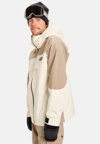 Beige og kremfarget skijakke med høy krage, glidelås-lommer og justerbare mansjetter. Brukt med en svart lue og skibriller.