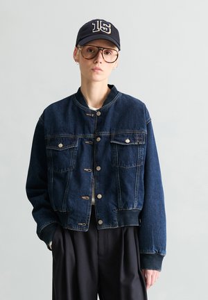 Person trägt eine dunkelblaue Denim-Bomberjacke, schwarz gerahmte Brille und eine marineblaue Mütze mit der Zahl 15, steht vor einem schlichten hellgrauen Hintergrund.