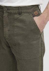 Pantalon cargo vert olive en mélange de coton, avec une fermeture à boutons, des poches latérales et un patch logo marron près de la taille.