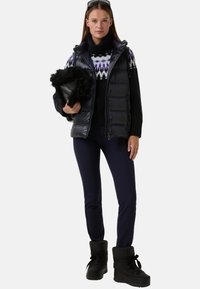 Lesklá černá puffer vesta s vysokým límcem, spárovaná s černým svetr s fialovým geometrickým vzorem, námořnické kalhoty a černé boty s kožešinou.