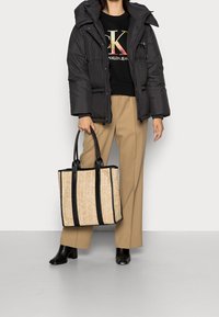 Svart puffjacka, svart tröja med logotyp, beige byxor, håller en tan- och svartrandig toteväska gjord av vävt material. Svarta klackstövlar.