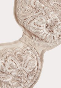 Soutien-gorge en dentelle beige clair avec motif floral, broderie délicate et texture de tissu lisse. Il présente des bonnets moulés et une bande sous poitrine douce.