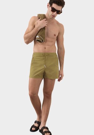 Mężczyzna bez koszulki, noszący oliwkowe kąpielówki, ciemne okulary przeciwsłoneczne, czarne sandały, trzyma złożony oliwkowy ręcznik na ramieniu, stoi na jednolitym tle.