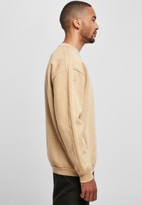 Sudadera beige con un ajuste relajado, puños acanalados y cuello redondo. Textura suave y diseño sencillo; detalles mínimos en la tela.