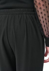 Zwarte broek met een elastische tailleband, met een gladde stoftextuur. Doorschijnende mouwen met zwart polkadotpatroon bij de manchetten.