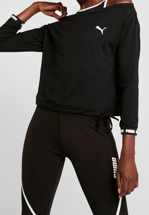 Personne portant un haut à manches longues noir Puma et des leggings noirs Puma, tirant légèrement le col du haut vers l'extérieur.