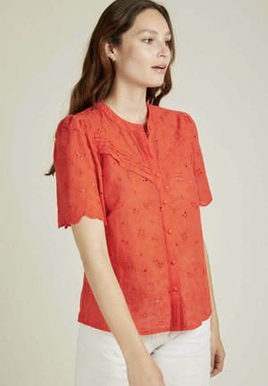 Blouse rouge brodée avec des manches courtes à bords festonnés, devant boutonné et motif floral. Tissu léger avec une apparence texturée.