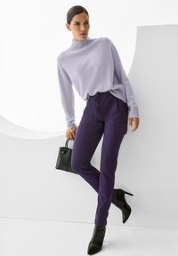 Vrouw in een lavendelkleurige trui en donkere paarse broek, met een zwarte handtas in haar hand, gekleed in zwarte enkellaarsjes met hak, staande tegen een witte muur.