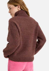 Suéter marrón de canalé con cuello alto, que presenta un tejido texturizado. La modelo lleva pantalones de pana rosa, visto desde la parte trasera.