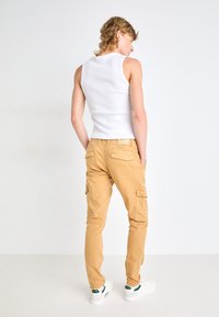 Tan cargo byxor med flera fickor, en åtsittande design, i bomullsmaterial, och en vit linne. Skorna är vita med gröna accenter.