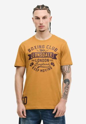 Joven con el cabello trenzado y tatuajes en las mangas, que lleva una camiseta amarilla mostaza con la inscripción "Lonsdale Boxing Club London" y vaqueros azules.
