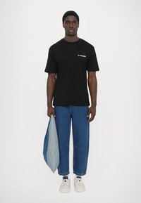 Μαύρο κοντομάνικο μπλουζάκι με τη λευκή επιγραφή "LES DEUX", συνδυασμένο με τζιν παντελόνι και λευκά αθλητικά παπούτσια, κρατώντας ένα ανοιχτό μπλε τζιν μπουφάν.