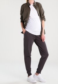 Zwangerschapsoutfit bestaande uit een wit T-shirt, een olijfgroen overhemd met knopen en donkergrijze joggingbroeken met enkelsluiting, gecombineerd met witte sneakers.