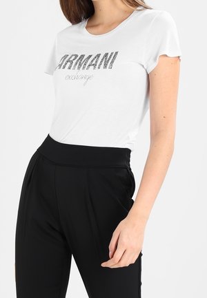 Frau trägt ein weißes Armani Exchange T-Shirt und hoch taillierte schwarze Hose, steht mit einer Hand nahe ihrer Hüfte.