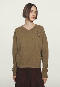 Donna con capelli corti e scuri che indossa un maglione a V in maglia marrone con una spilla astratta argento e pantaloni scuri su uno sfondo neutro.