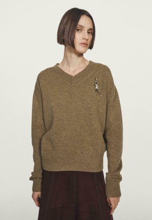 Donna con capelli corti e scuri che indossa un maglione a V in maglia marrone con una spilla astratta argento e pantaloni scuri su uno sfondo neutro.
