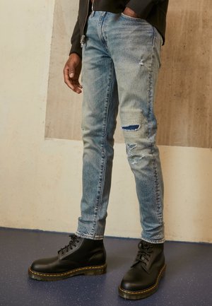 Personne portant un jean déchiré bleu clair et des bottes noires à lacets, debout contre un mur beige et blanc.