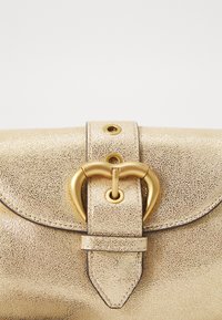 Goldfarbene Handtasche mit metallischer Textur, ausgestattet mit einem herzförmigen goldenen Verschluss und einem Tragegurt, der mit goldenen Nieten gesichert ist.