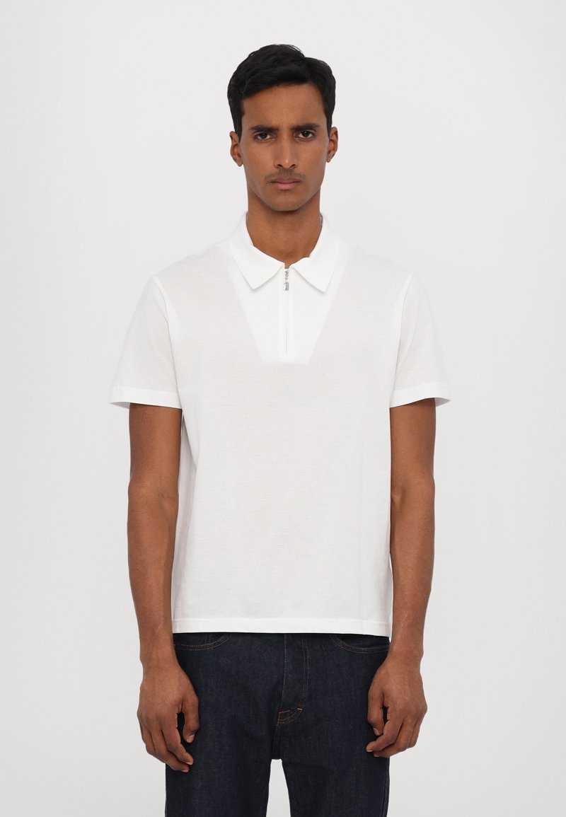 Balta polo marškinėliai su užtrauktukų apykakle, trumpomis rankovėmis ir tekstūruotu priekinio skydelio paviršiumi. Sukurti su laisvu siluetu ir minimalistiniu stiliumi.