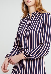 Košilové šaty s knoflíky v námořnické modré, růžové a bílé pruhy. Mají dlouhé rukávy s elastickými man žetami, páskem v oblasti pasu a klasický límec.