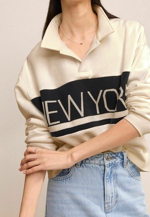 Femme portant un sweat-shirt crème avec l'inscription noire "NEW YORK" et un jean bleu clair, les bras croisés, et un collier argenté avec un pendentif rouge.