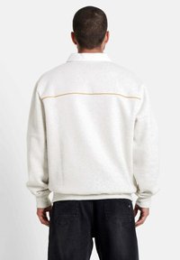 Lichtgrijze, oversized sweatshirt met een witte kraag, voorzien van een contrasterende gele naad over de rug. Zachte stof, lange mouwen.