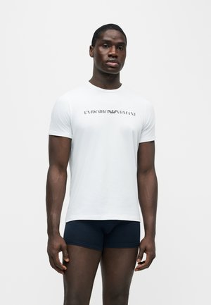 Mann trägt ein weißes Emporio Armani Logo-T-Shirt und dunkle, enganliegende Shorts und steht vor einem schlichten hellen Hintergrund.