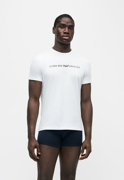 Άνδρας που φοράει λευκό T-shirt με το λογότυπο Emporio Armani και σκούρα εφαρμοστά σορτς, στέκεται μπροστά σε απλό ανοιχτό φόντο.