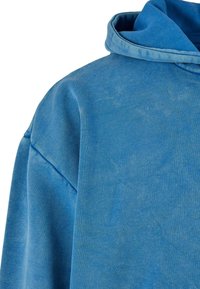 Sweat à capuche bleu en tissu texturé et doux. Il présente une grande capuche et des épaules tombantes, avec une coupe décontractée et des variations de couleur subtiles.