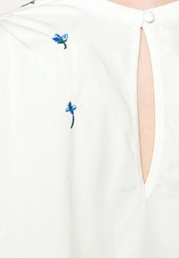White cotton blouse featuring blue floral embroidery and a keyhole opening at the back, accented with a button closure.