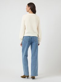 Mulher de costas, vestindo um suéter branco felpudo, jeans retos azul claro e botas de cano curto castanhas com salto, em fundo simples.