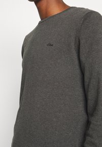 Pull en maille gris avec un col rond et un petit emblème sur la poitrine. La texture est douce et de poids moyen.