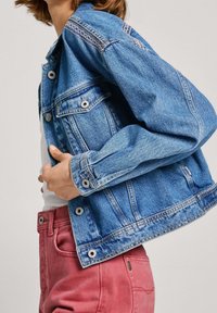 Jasnoniebieska kurtka dżinsowa z dwiema przednimi kieszeniami, srebrnymi guzikami i przeszyciami na ramionach, w zestawieniu z różowymi spodniami.