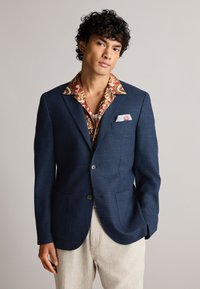 Blazer ajusté bleu marine à deux boutons, en tissu texturé, avec deux poches à l'avant, associé à une chemise à motifs et un pantalon beige clair.