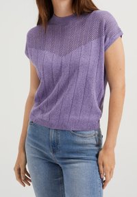 Kvinna iklädd en kortärmad lila stickad topp med sicksack- och vertikala randmönster, tillsammans med ljusblå denimjeans.